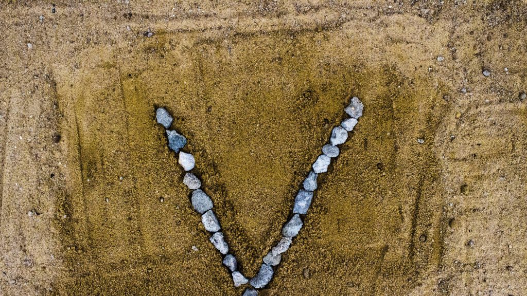 Minimalist letter V made of gray stones on textured sandy brown surface nature wallpaper background