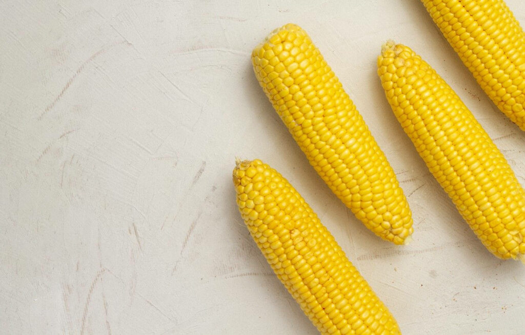 Minimalistic Wallpaper: Four Yellow Corn Cobs on White Table Surface