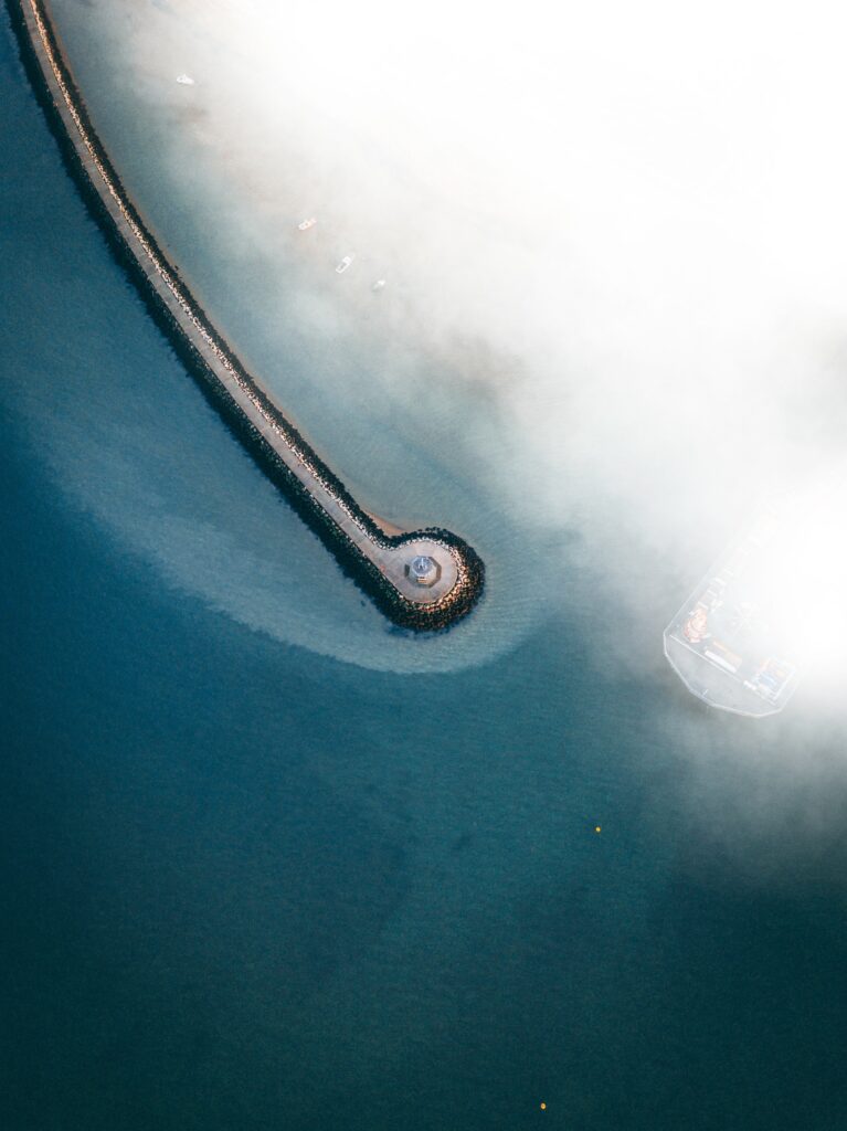 Aerial view of a curving pier over calm blue sea with mist creating a serene coastal scene Wallpaper
