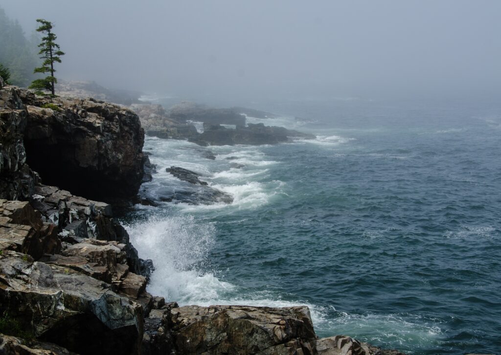 Misty cliffside with rugged rocks and foamy ocean surf under a tranquil blue sky wallpaper