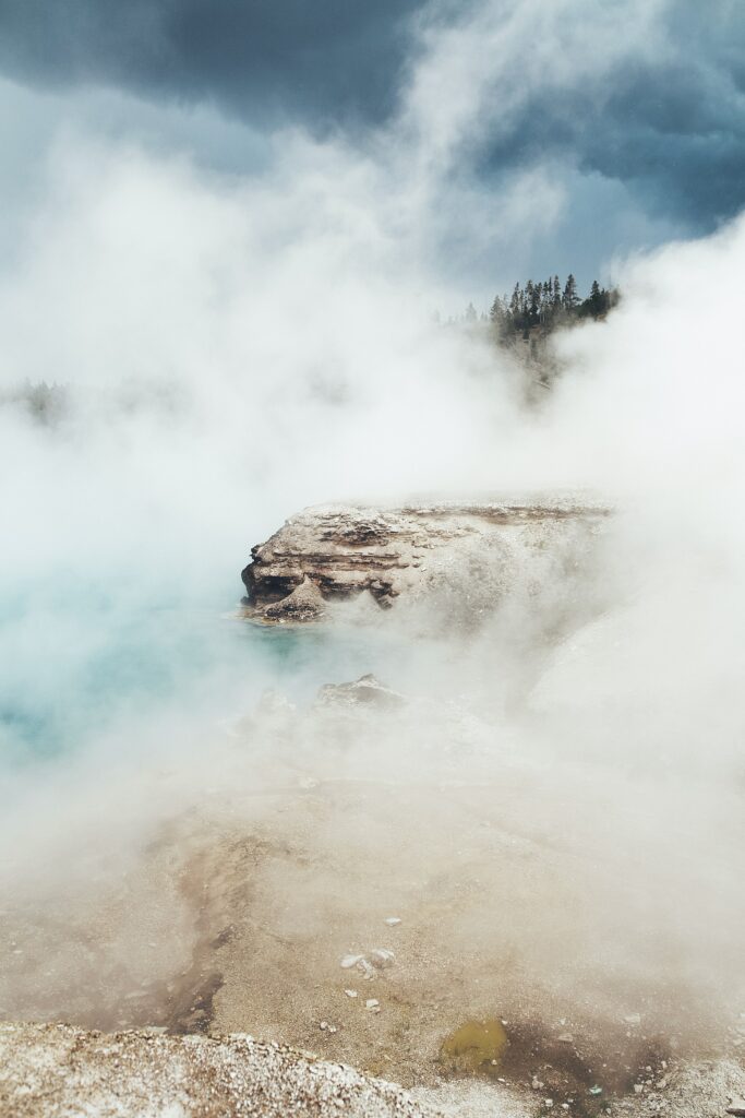 Misty coastal rocks with fog swirling near towering trees under moody sky over pale blue water Wallpaper