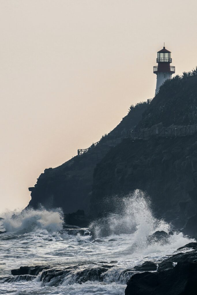 Solitary lighthouse on rugged rocks with crashing waves under misty sky coastal landscape wallpaper