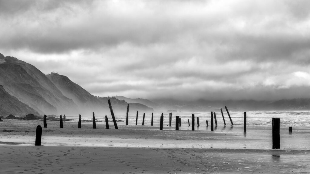 Black and white coastal wallpaper with weathered pilings misty shoreline rugged mountains and clouds