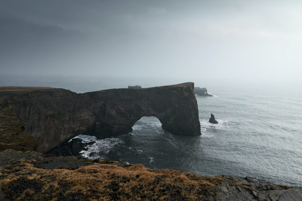 Dramatic stone arch over misty sea with crashing waves and golden grass coastal landscape wallpaper