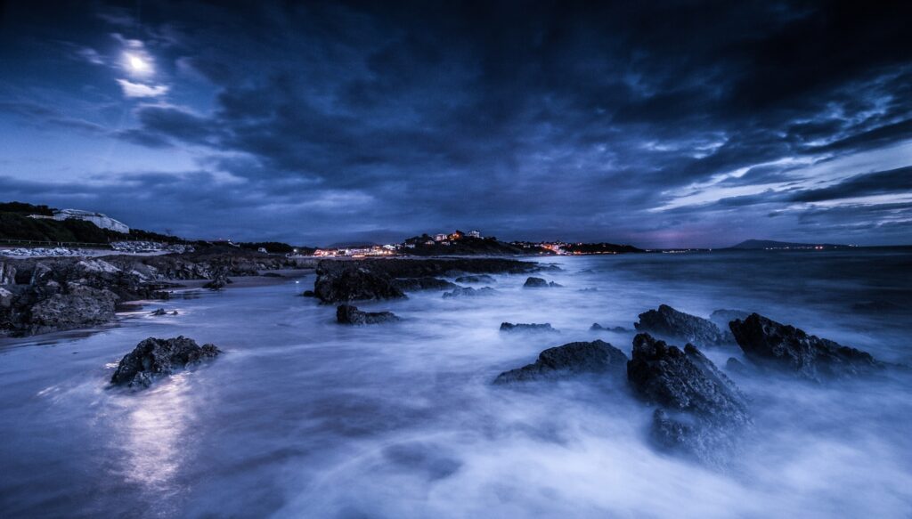 Dramatic night seascape with moonlit waves crashing on rocks and distant city lights wallpaper
