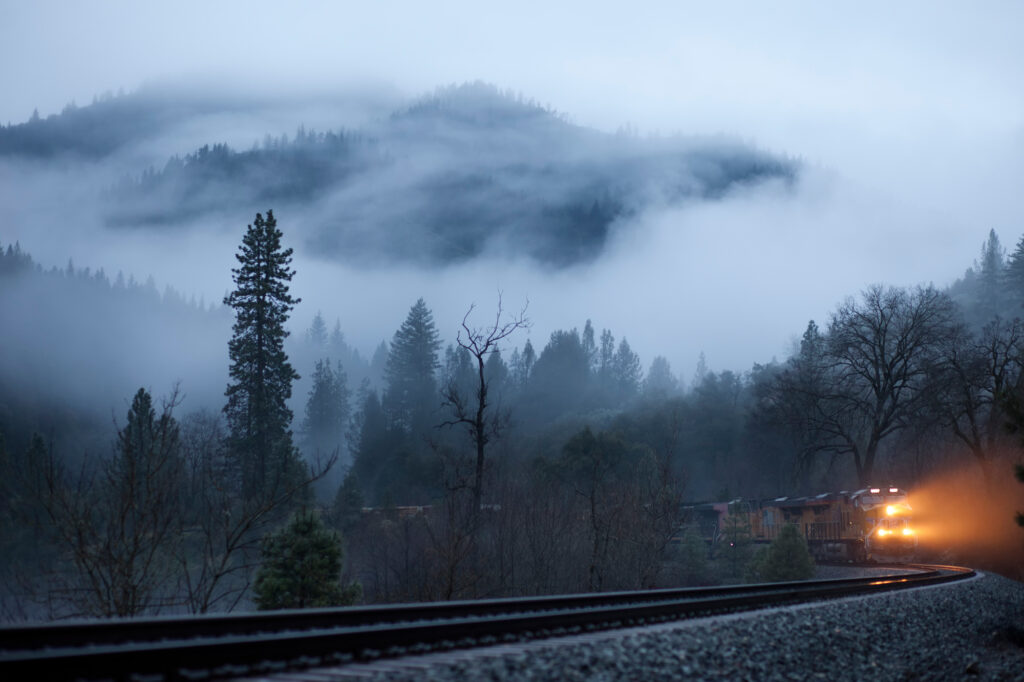 Train with bright headlights moving on curved rails through foggy forested mountains in morning Wallpaper