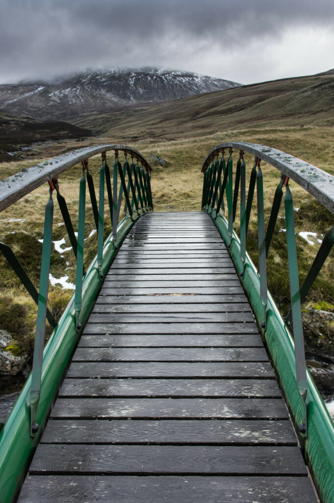 Narrow green bridge over misty stream leading to foggy snow-dusted mountains and gray sky Wallpaper