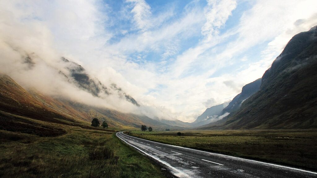 Winding wet asphalt road through misty mountain valley with dramatic cloudy sky and peaks Wallpaper