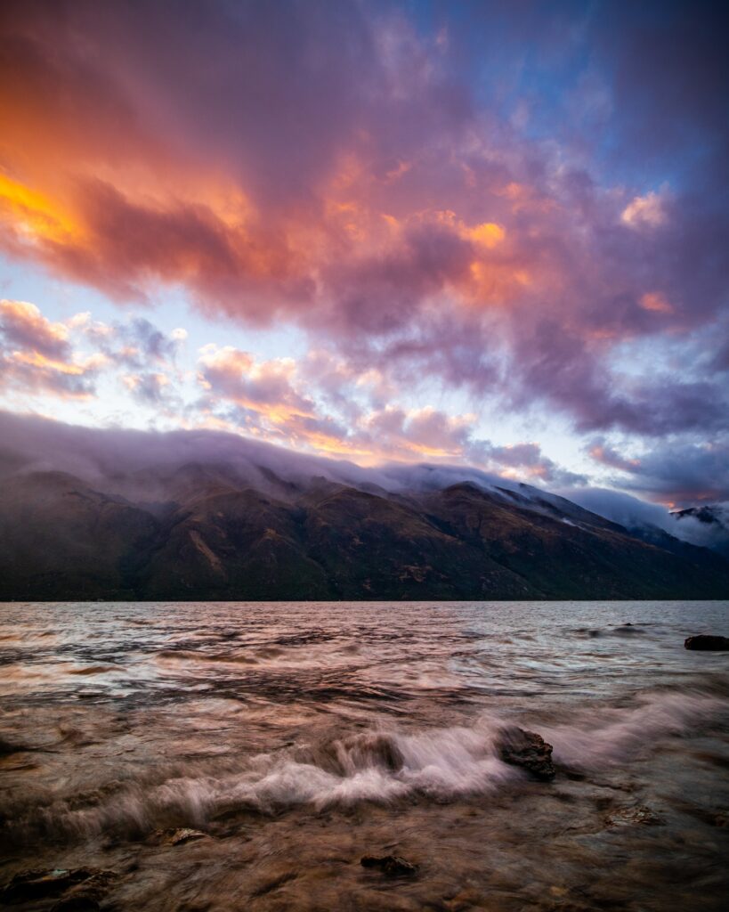 Vibrant river shoreline with rocky edges misty foggy hills and colorful dramatic sunset clouds wallpaper