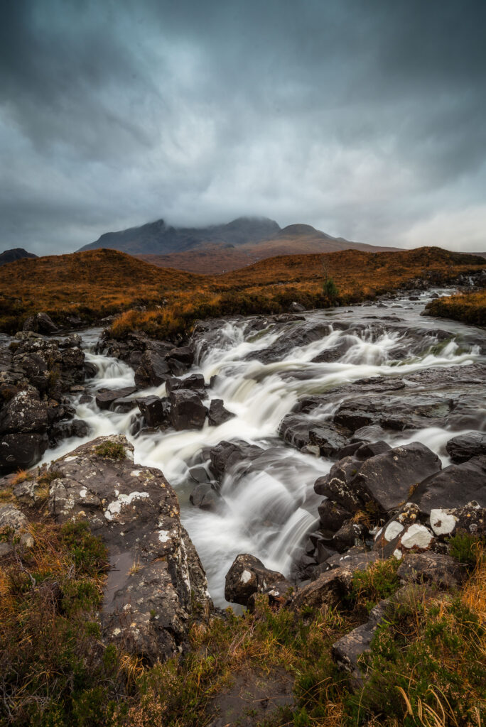 Misty river flows over rugged stones and waterfall with golden grass foggy mountains dreamy sky Wallpaper