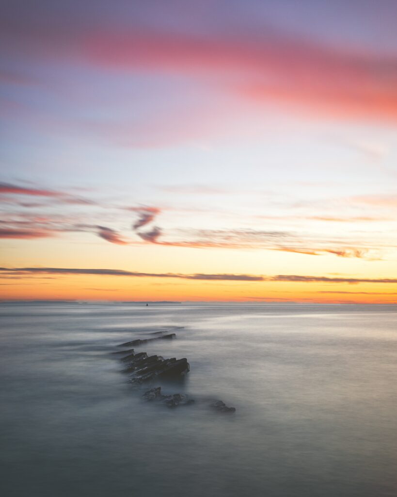 Misty rocks in calm waters at dusk with pastel pink orange clouds and soft foggy sky background Wallpaper