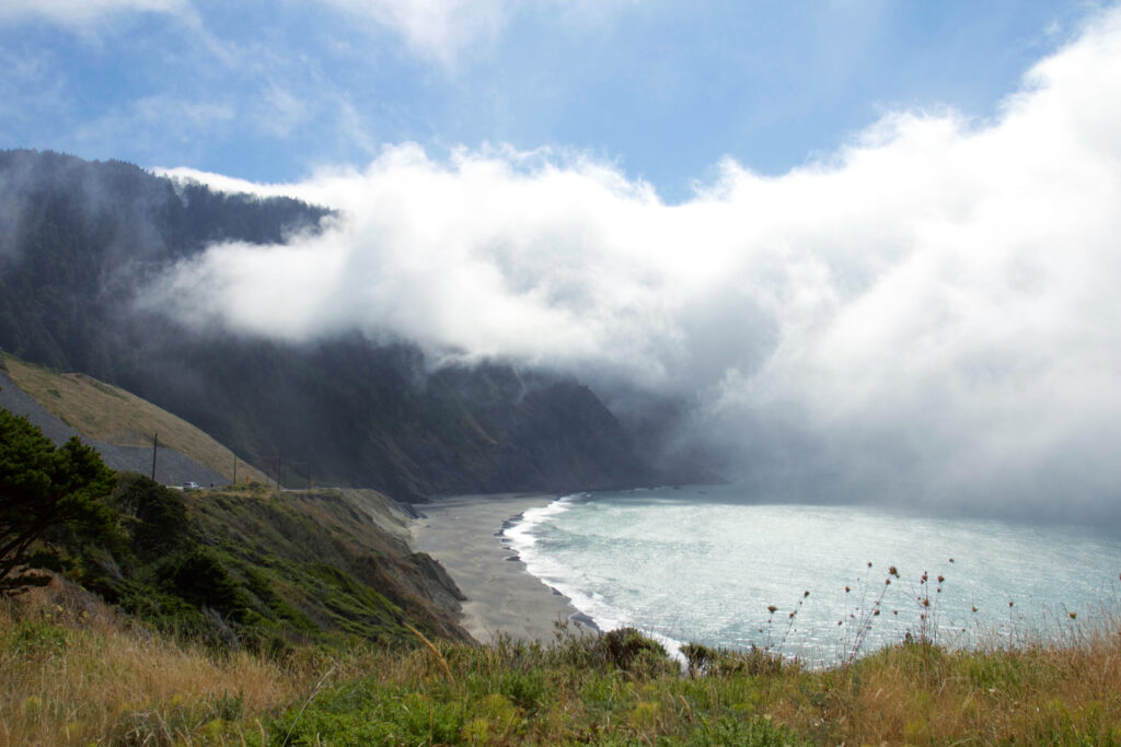 Mist-covered shoreline with gentle waves sandy beach rugged mountain and grass-covered cliffs Wallpaper