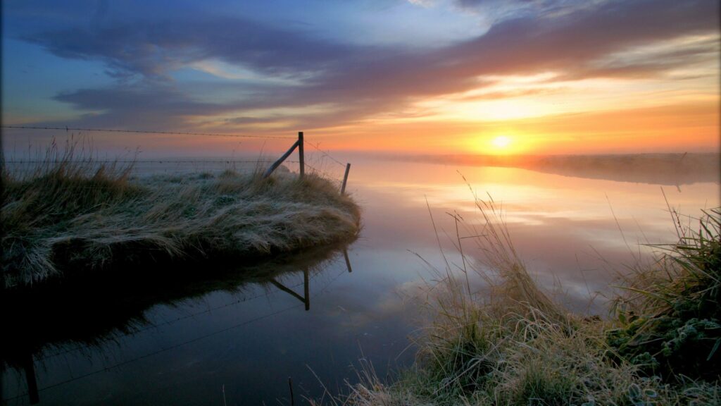 Misty sunrise over calm coastal water with frosted grass and rustic fence under dramatic sky Wallpaper