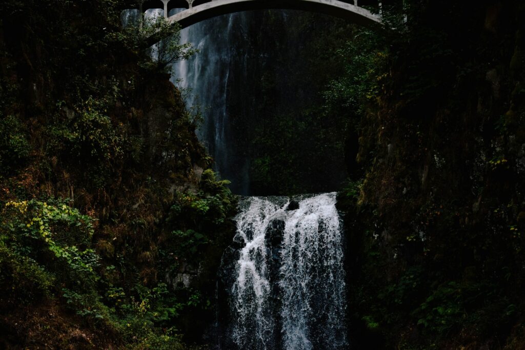 Dramatic waterfall under stone bridge with lush green trees and misty forest background wallpaper