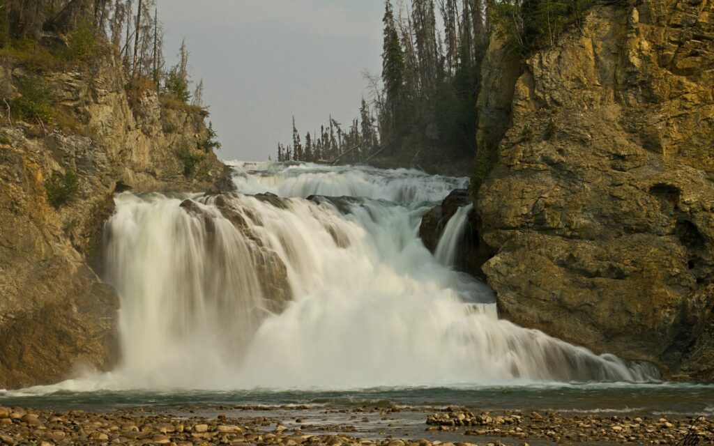 Majestic waterfall over rocky cliffs flows into stream with sparse trees under cloudy sky wallpaper