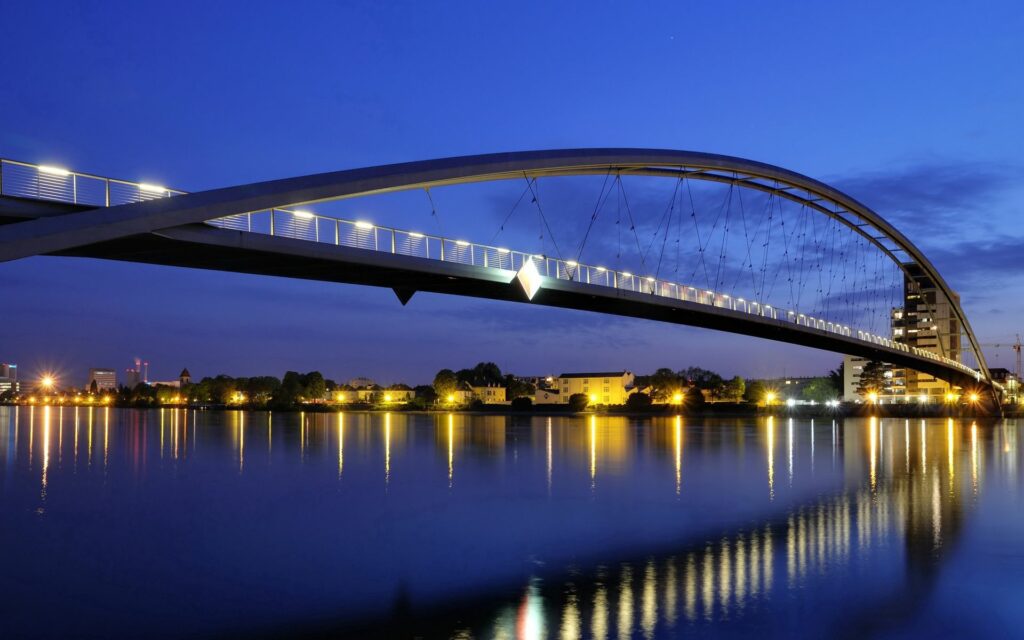 Modern arch bridge glowing at twilight reflecting on calm river with warm city lights background Wallpaper