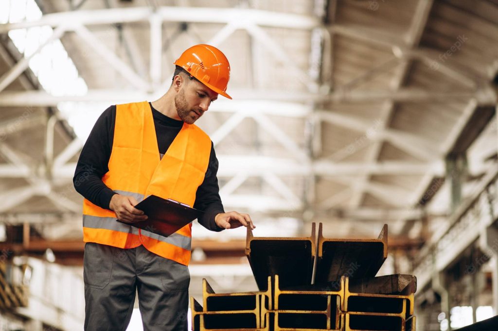 Construction worker in orange vest inspects steel I-beams inside bright industrial warehouse interior Wallpaper