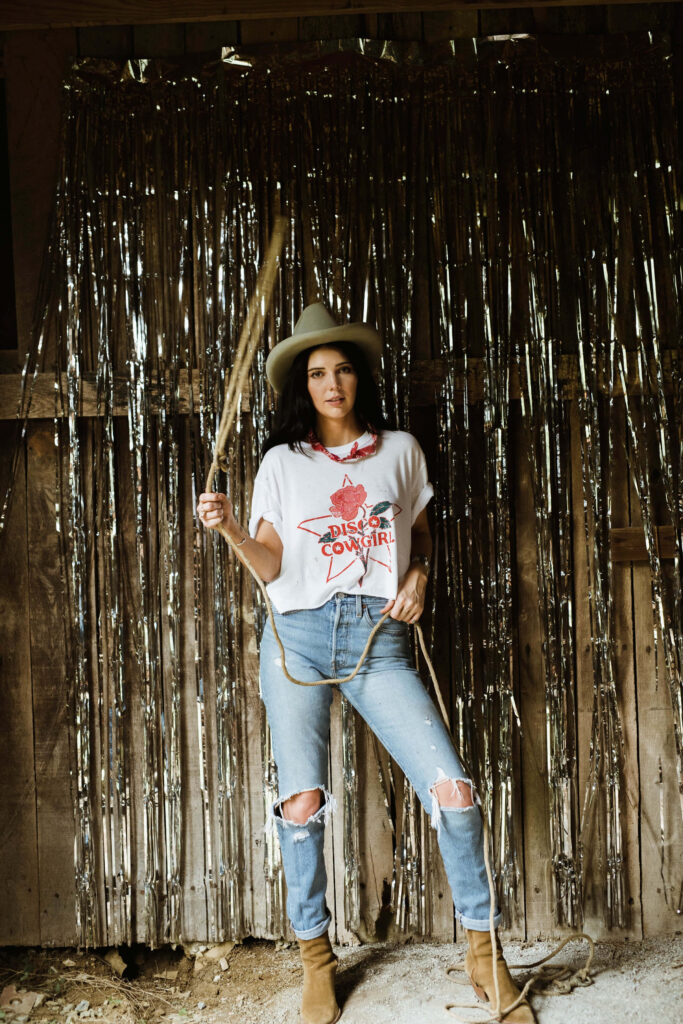 Stylish cowgirl with lasso in rustic barn against shimmering metallic streamers background wallpaper