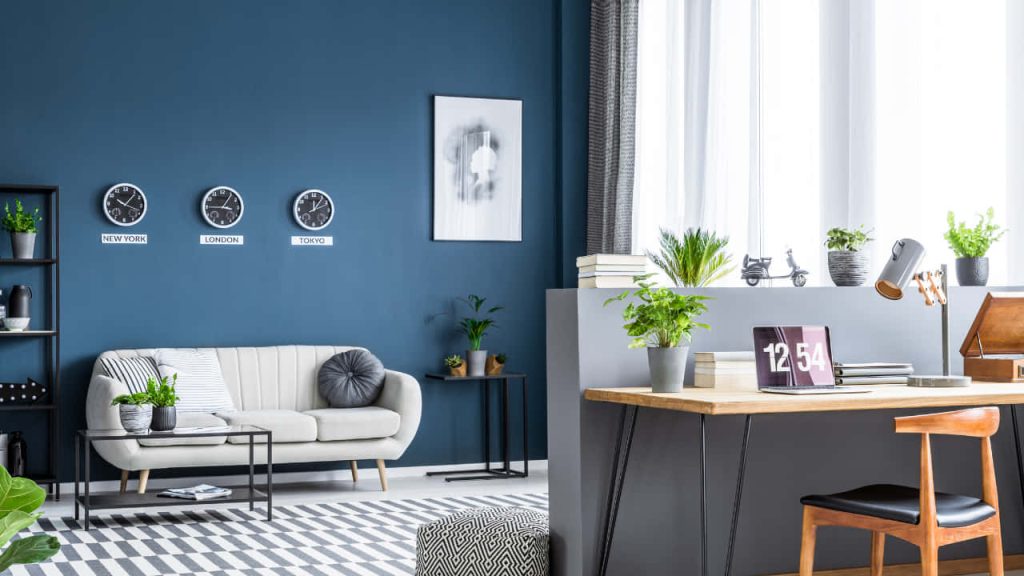 Modern home office Zoom background with deep blue walls gray couch wooden desk and clocks Wallpaper