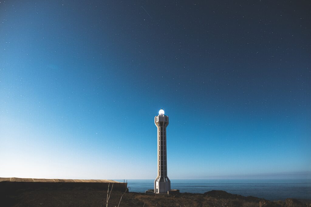 Tall modern white lighthouse on rugged coast under clear starry night sky serene seaside scene Wallpaper
