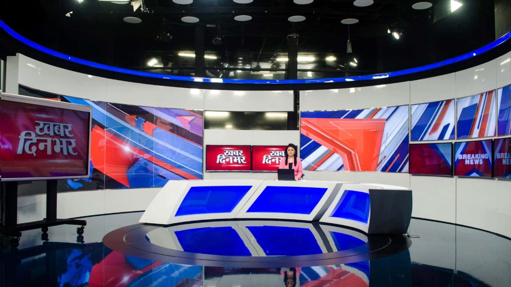 News anchor in pink blazer at blue-lit desk with vibrant geometric Hindi news studio background Wallpaper