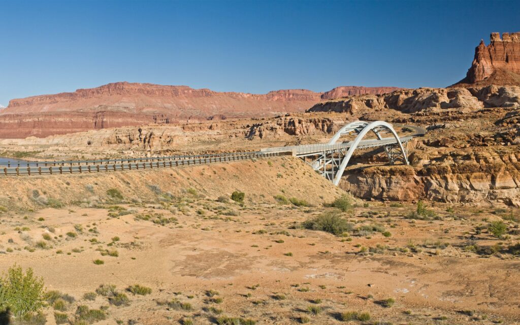 Modern white bridge curving over rugged desert canyon with red rocks and clear blue sky wallpaper