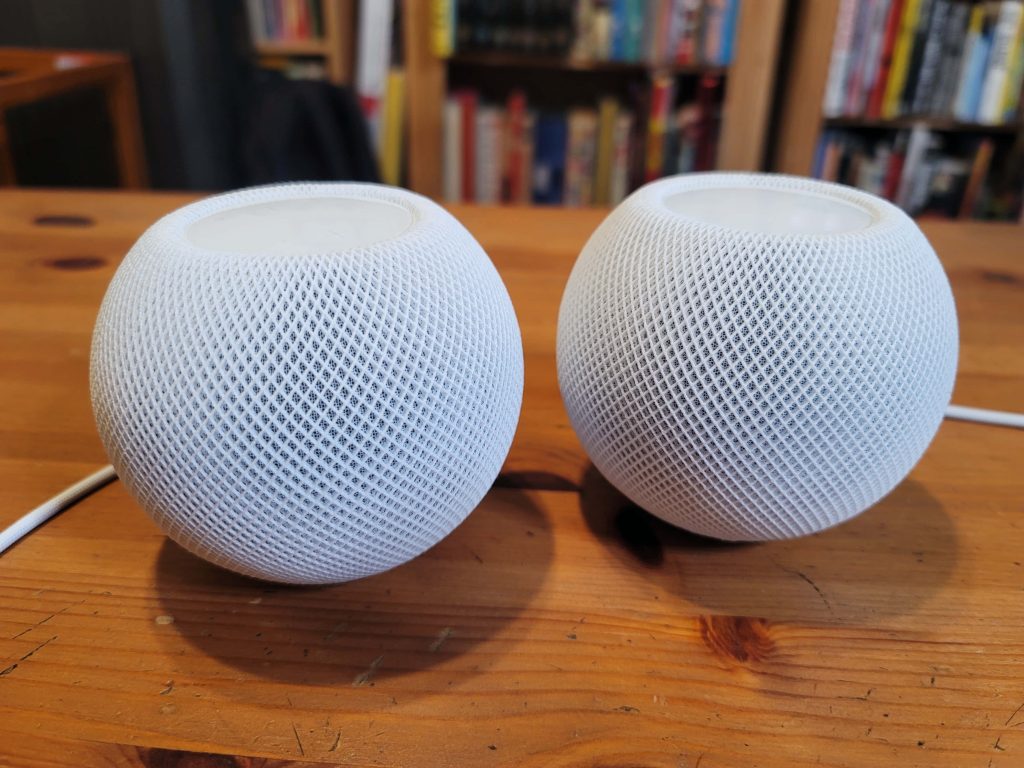 Two white Apple HomePod mini speakers on wooden desk with blurred bookshelf background wallpaper