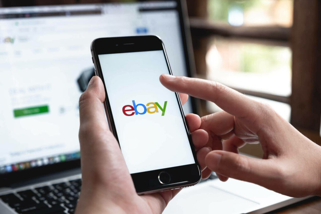 Person holding smartphone showing colorful eBay logo with blurred laptop in modern ecommerce scene Wallpaper