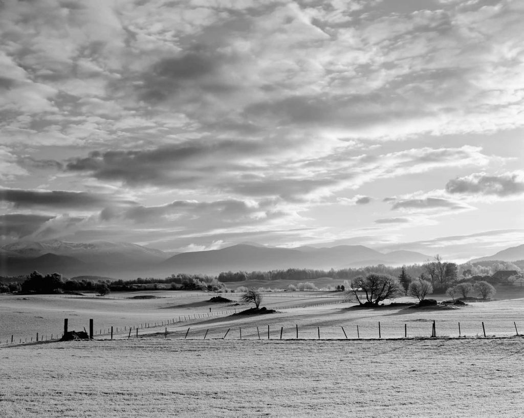 Monochrome landscape with dramatic clouds frosty fields wire fences and distant mountains Wallpaper
