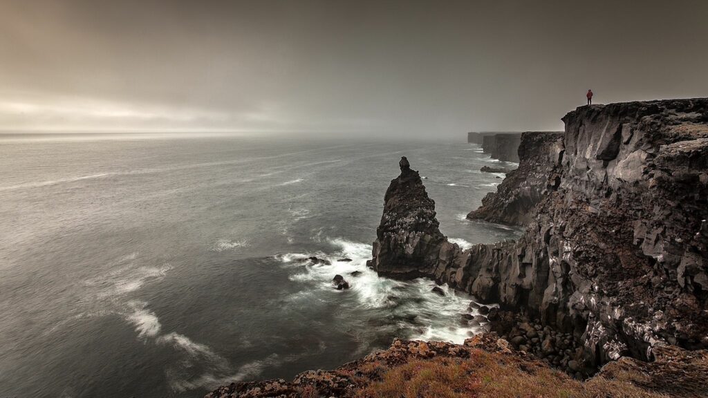 Dramatic coastal cliffs with misty sea rocky formations and lone figure under moody sky wallpaper