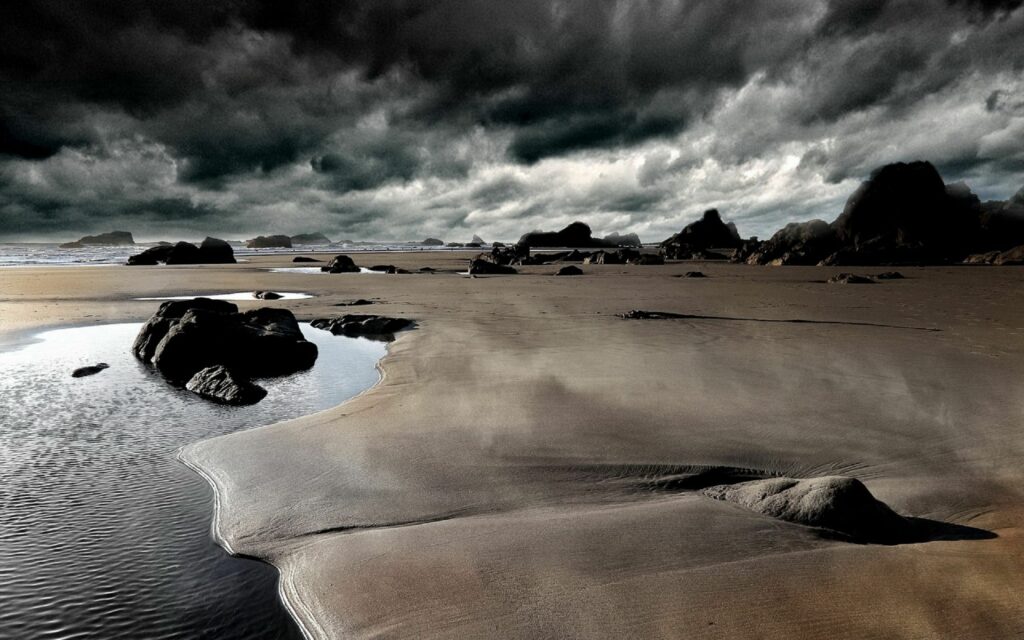 Dramatic coastal wallpaper with smooth sand dark rocks and reflective pools under cloudy sky