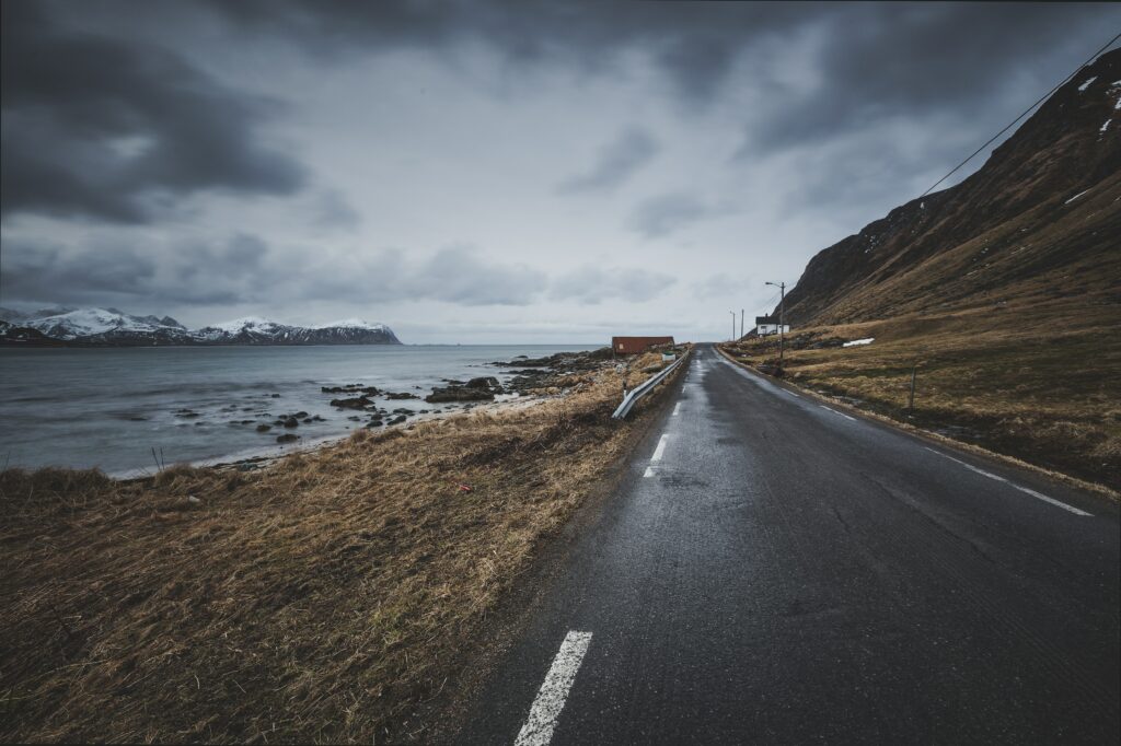 Narrow coastal road beside windswept grass and rugged mountains under dark cloudy sky wallpaper