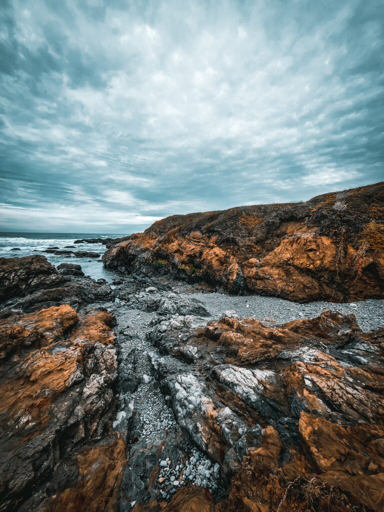 Moody coastal landscape with rugged orange rocks and dramatic overcast sky over blue water Wallpaper