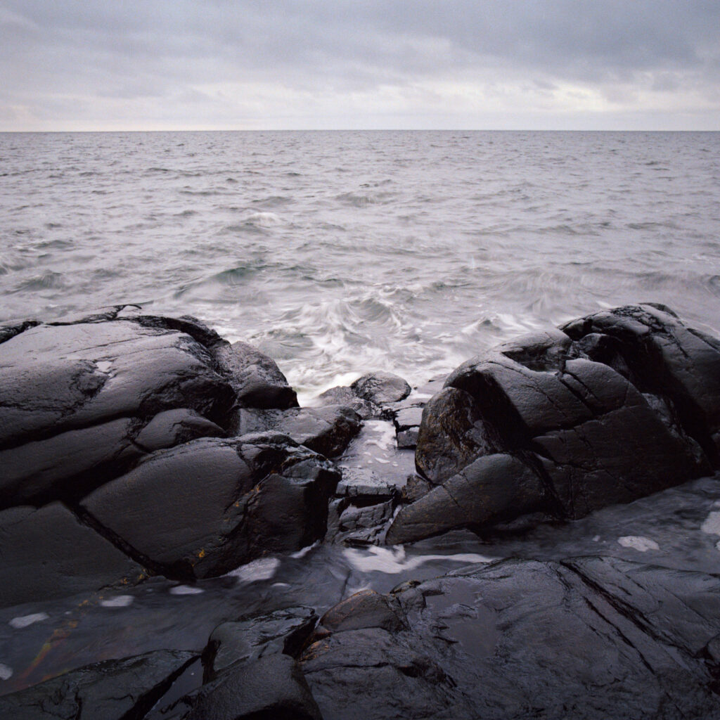 Dark rugged coastal stones and boulders framing a serene sea below cloudy moody sky wallpaper