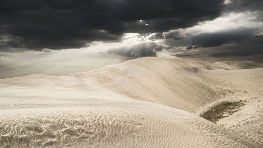 Stormy sky over rippled beige sand dunes desert landscape wallpaper with soft light beams