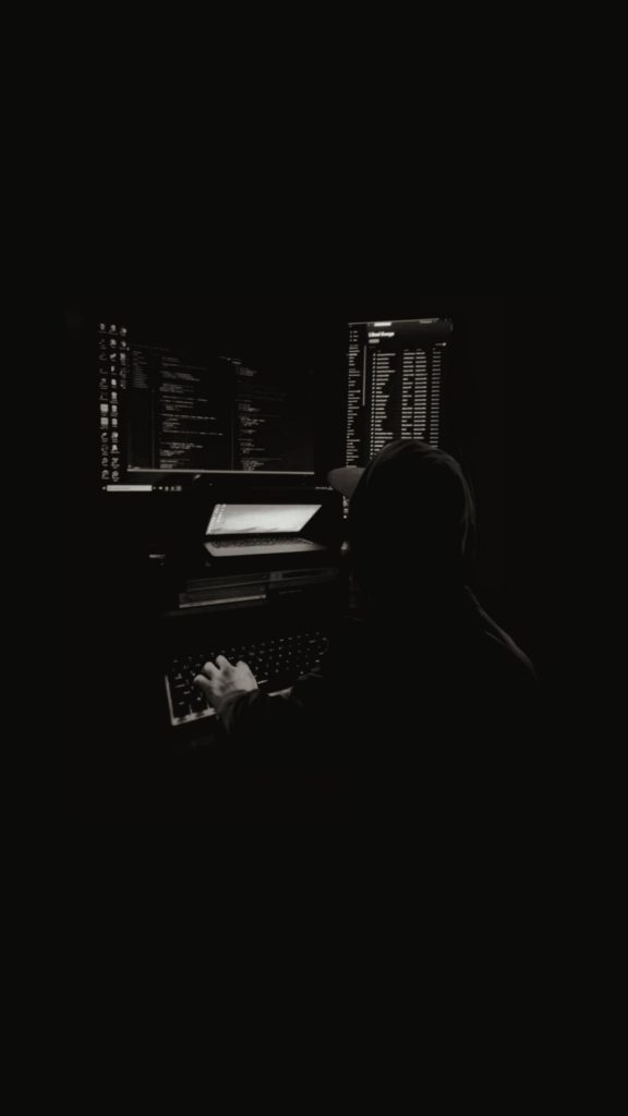 Moody grayscale wallpaper of hooded hacker coding at dark workstation for tech phones