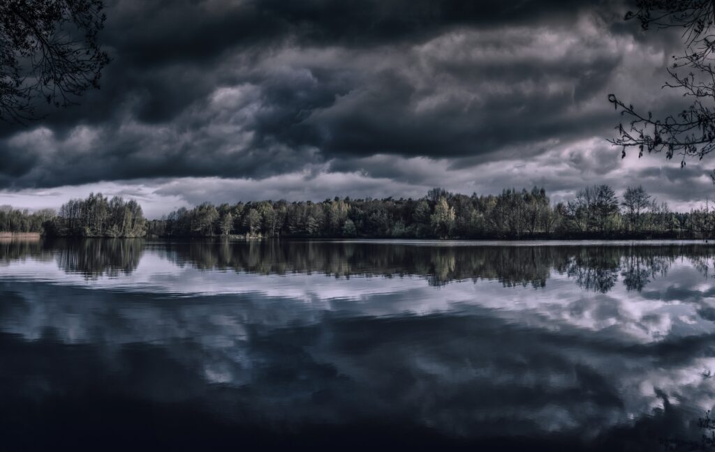 Moody lake landscape with dense trees and dark clouds reflecting on still water wallpaper background