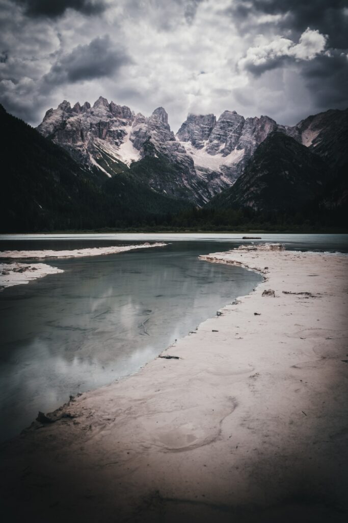 Rugged mountain peaks reflected in calm lake under cloudy sky with sandy shore foreground Wallpaper