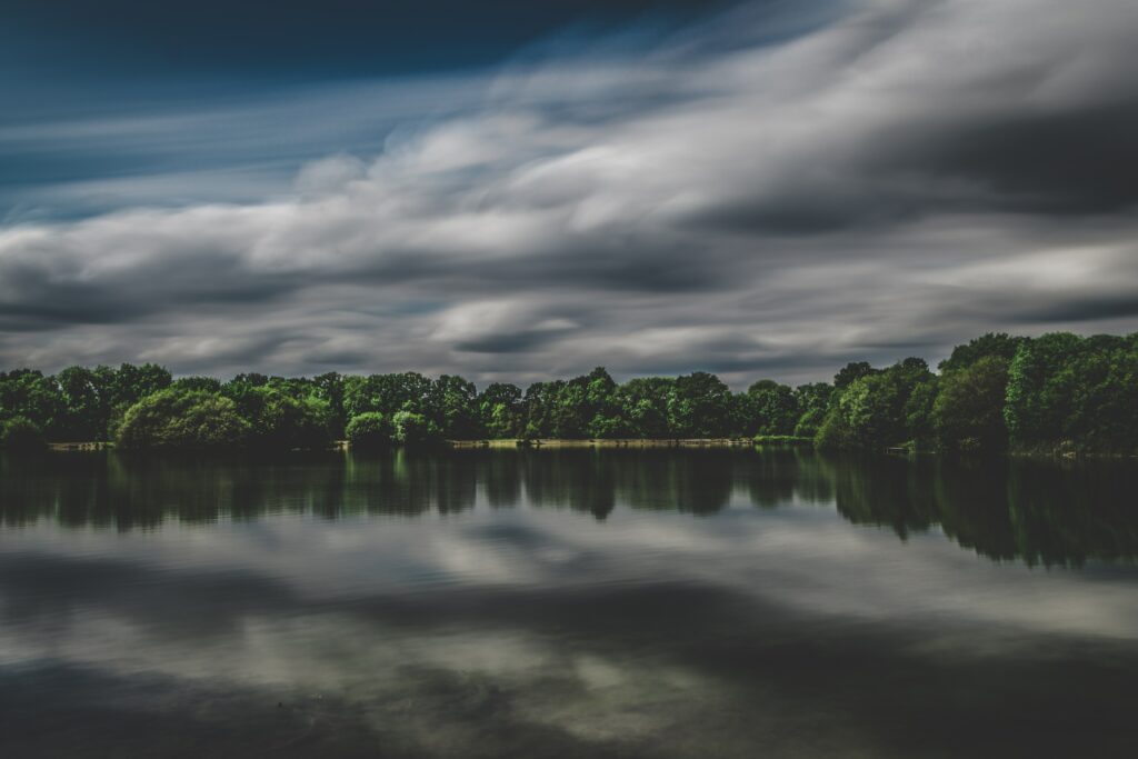 Serene lake with lush green trees reflected in still water under dramatic overcast sky wallpaper background