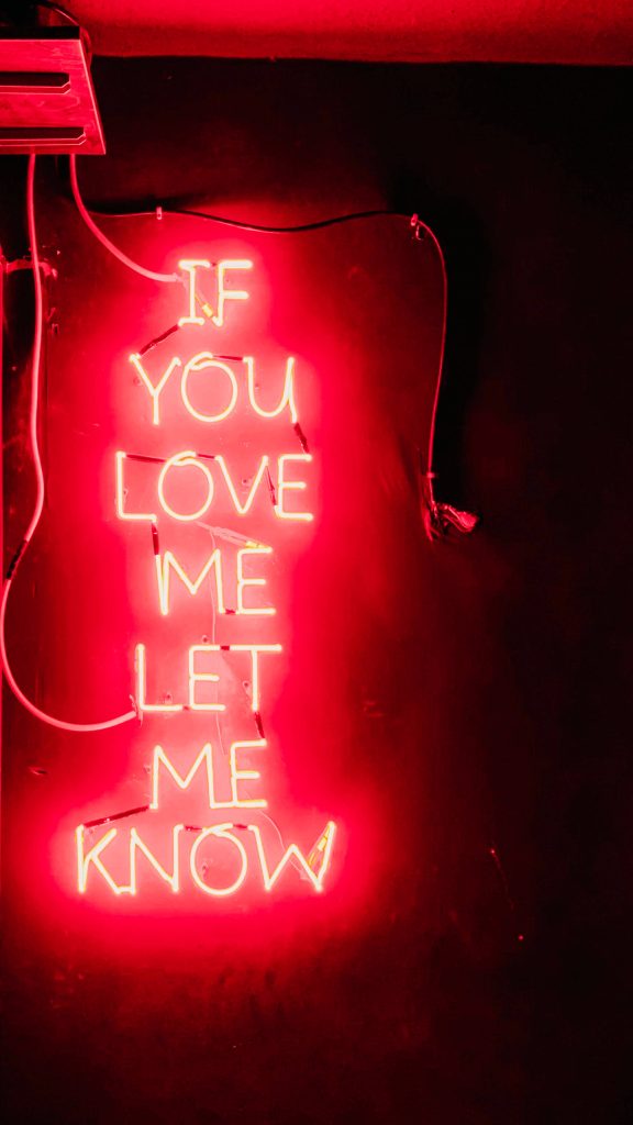 Red neon sign saying If You Love Me Let Me Know on dark textured wall iPhone wallpaper background