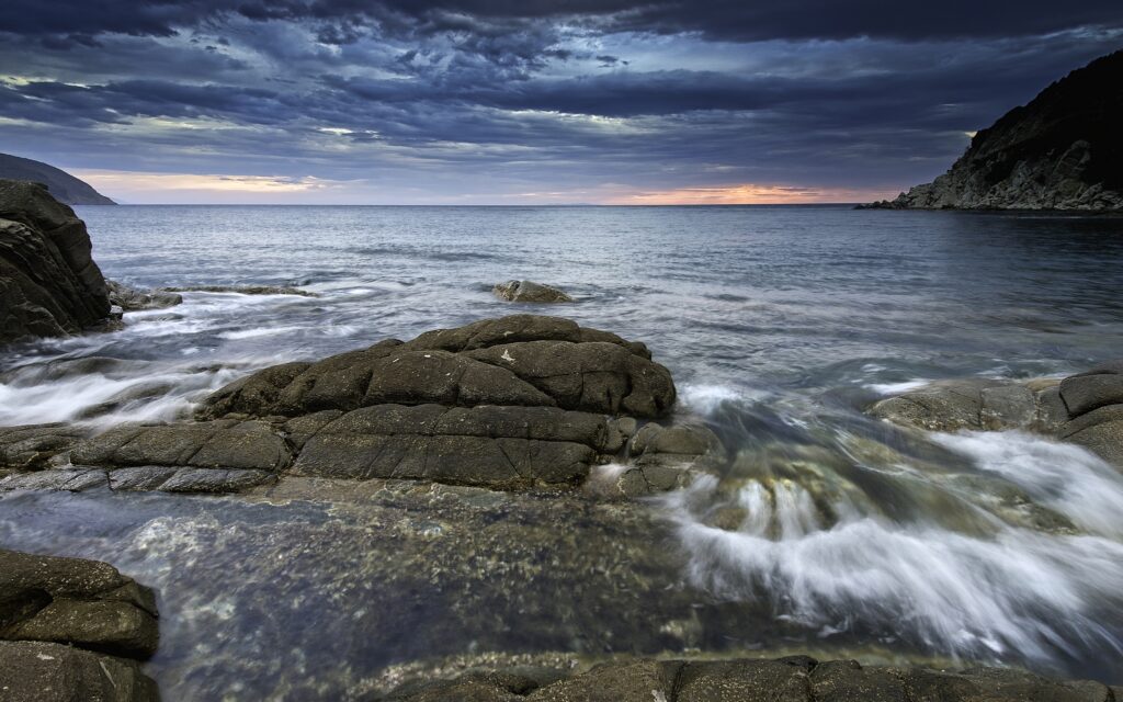 Dramatic seascape with dark rocks gentle waves serene bay moody clouds and sunset glow wallpaper