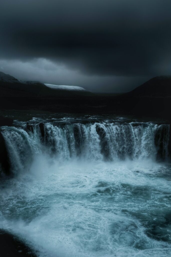 Dramatic waterfall flowing into mist under dark sky with moody blue and gray tones background Wallpaper