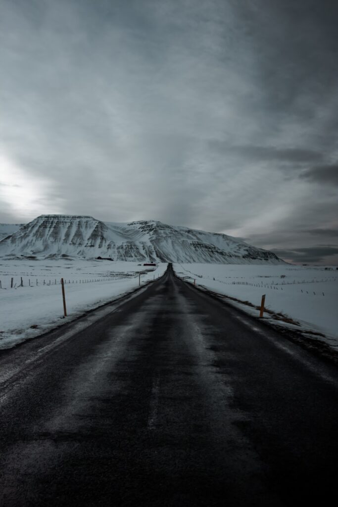 Wet long road through snowy fields and rugged mountains under dramatic overcast winter sky wallpaper