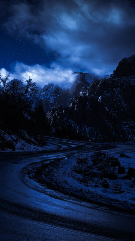 Moonlit winding mountain road with snow and rugged peaks under starry night sky wallpaper