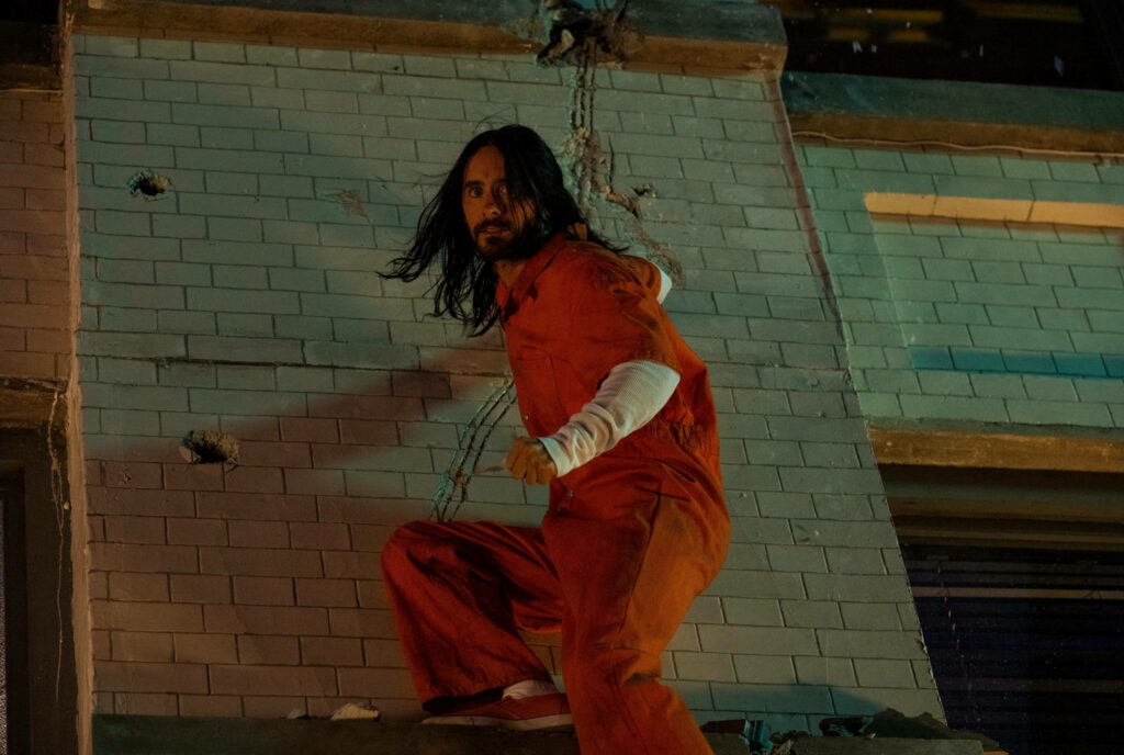 Morbius in orange prison uniform crouching by tiled wall with chains creating intense scene Wallpaper