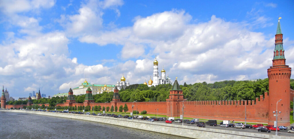 Moscow Kremlin landscape with red brick walls golden domes and riverbank greenery under blue sky Wallpaper