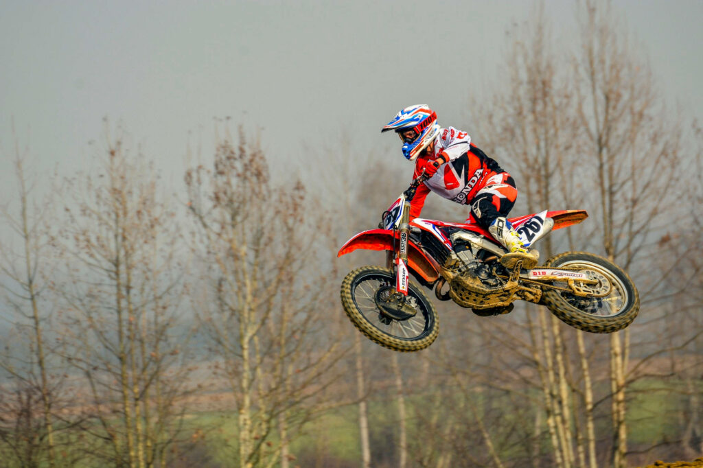 Dirt bike rider in red and white gear mid-air jump over rugged landscape with cloudy sky Wallpaper