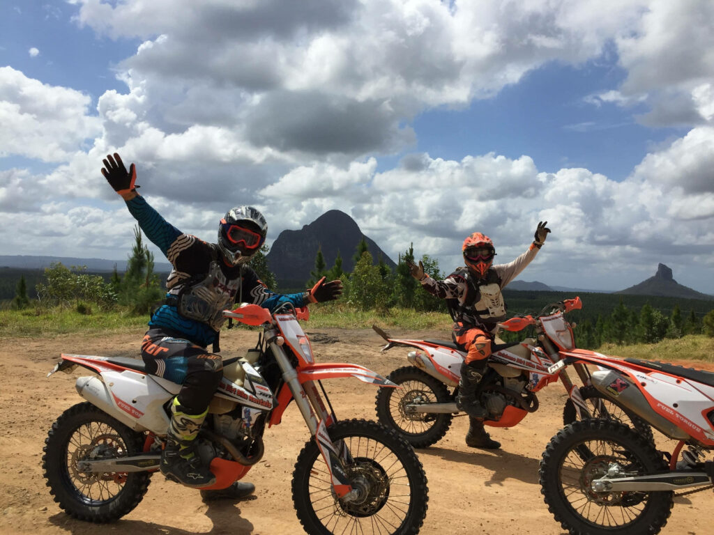 Two dirt bike riders in full gear celebrating on rugged terrain with mountains and cloudy sky background Wallpaper
