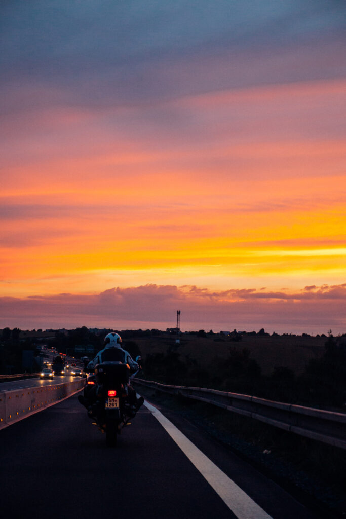 Motorcyclist riding open road at vibrant orange pink purple sunset sky with countryside backdrop Wallpaper