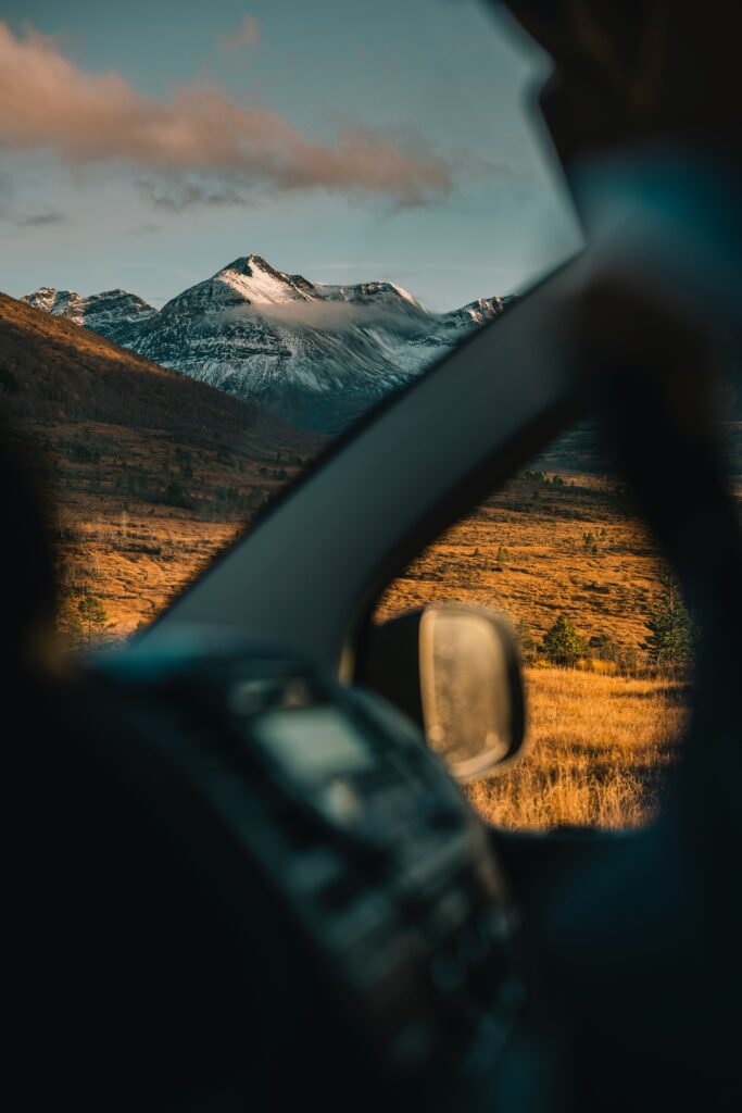 Snow-capped mountain landscape viewed from car window with golden fields and warm sunlight Wallpaper